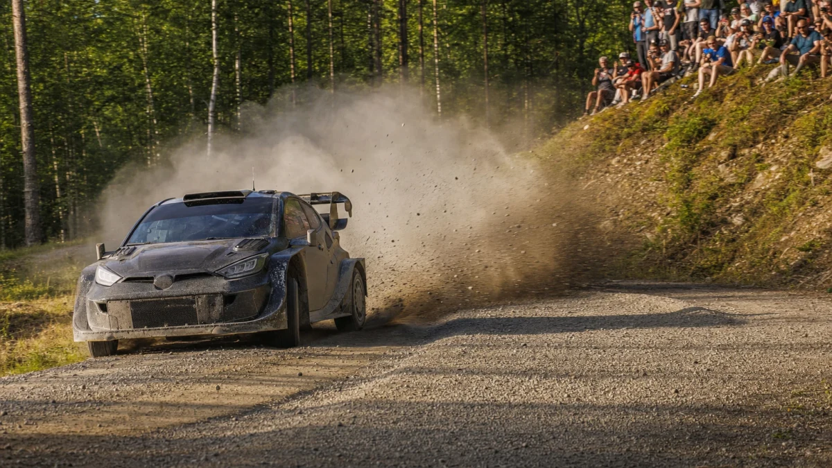 Rallybil som kastar upp grus och damm genom en kurva på en grusväg i skogen, med åskådare på slänten bakom.